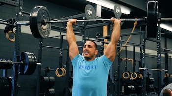 Un hombre con camiseta azul levanta una barra con discos negros por encima de la cabeza en un gimnasio