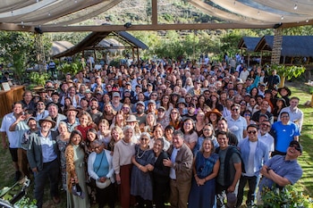 Foto grupal de una gran multitud de personas al aire libre, muchas sonrientes, bajo un toldo, con árboles y cabañas de techo de paja al fondo