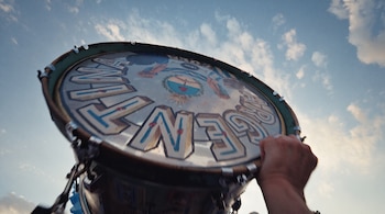 Una persona sostiene un bombo con la palabra "Argentina" y el Escudo Nacional grabados en su parche, contra un fondo de cielo azul con nubes blancas.