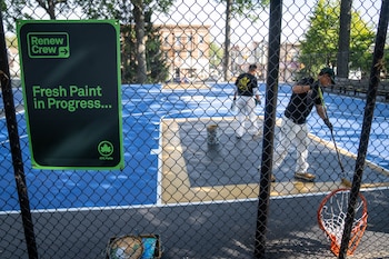 Un cartel de Renew Crew fijado en una cerca metálica ante dos trabajadores que pintan el suelo de una cancha deportiva