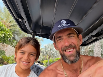 Un hombre barbudo con gorra y torso descubierto, y un niño sonriente, sentados en un carrito de golf. Detrás se observan palmeras