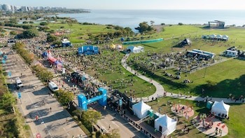 Vista aérea de un gran evento al aire libre en un parque verde junto al río. Miles de personas transitan una carretera y áreas de césped con carpas, arcos y un skyline lejano