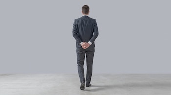 Un hombre de traje oscuro y camisa blanca camina de espaldas, con las manos entrelazadas detrás de la espalda, sobre un suelo de concreto gris claro.