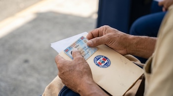 Primer plano de manos adultas de tez trigueña sosteniendo una cédula de identidad costarricense y una carpeta beige con el logo de Migración y Extranjería.