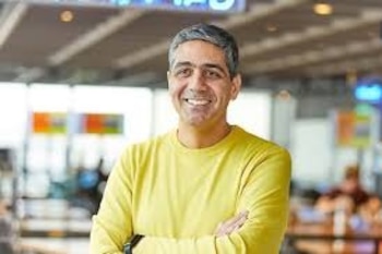 Hombre de mediana edad con cabello grisáceo y camiseta amarilla sonríe con los brazos cruzados. Fondo de interior borroso con luces y personas