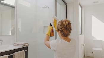 Una persona con guantes amarillos aplica un producto sobre una mampara de cristal con un paño blanco en un baño luminoso.