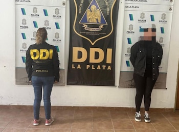 Dos mujeres de pie frente a banners de la Policía DDI La Plata, una con chaqueta policial amarilla y la otra de ropa oscura con el rostro pixelado