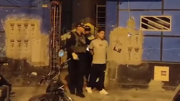Un hombre esposado avanza junto a dos policías en la vía pública frente a una fachada iluminada durante la noche