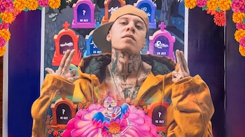 Hombre con tatuajes, gorra y chamarra mostaza sobre fondo de lápidas de colores, flores de cempasúchil y una calavera con vestido rosa.