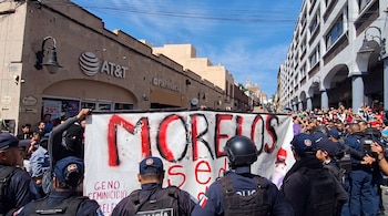 Elementos de policía y manifestantes frente a una gran pancarta blanca con letras rojas en una calle con comercios y edificios.