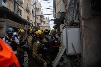 Grupo de hombres con uniformes, cascos (amarillos y azules) y chalecos tácticos camina por una calle estrecha con escombros. Se ven edificios y cables
