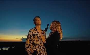 Hombre joven con camisa estampada y mujer rubia con arma, apuntando hacia él, bajo un cielo crepuscular azul y naranja con un paisaje oscuro de fondo