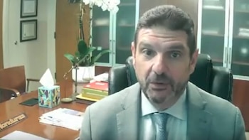 Hombre de barba con traje gris y corbata azul en videollamada; fondo de oficina con orquídea blanca, caja de pañuelos y documentos