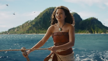 Catherine Laga'aia como Moana en un bote. Viste un top y falda marrones, un collar turquesa, y sonríe mientras sujeta una cuerda con montañas de fondo