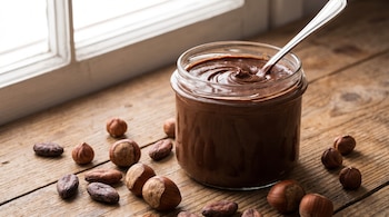 Tarro de cristal con crema de cacao y avellanas, una cuchara metálica. Avellanas enteras y granos de cacao sobre mesa de madera rústica.