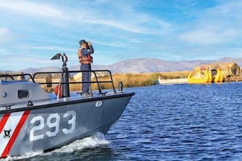 Un oficial con chaleco salvavidas mira por binoculares desde una lancha gris de la Marina de Guerra (N° 293) en el Lago Titicaca, con islas de totora y montañas al fondo