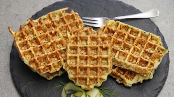 Cuatro waffles dorados con trozos verdes sobre un plato de pizarra negra, un tenedor de metal y tiras de calabacín.