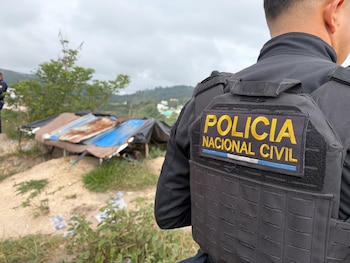 Espalda de un agente policial con chaleco táctico negro y parche "Policía Nacional Civil". Fondo con estructuras improvisadas de chapa y lona en terreno irregular