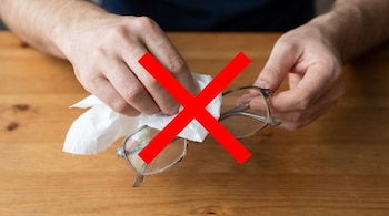 Primer plano de manos sosteniendo unas gafas sucias y un trozo de papel higiénico. Una gran cruz roja se superpone a la escena, indicando una acción incorrecta.