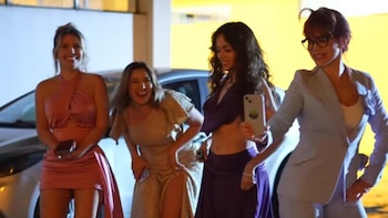 Cuatro mujeres sonrientes en vestidos de noche y un traje posan frente a un coche plateado de noche, con una de ellas sosteniendo un teléfono para una selfie