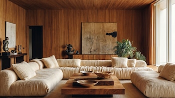 Una sala de estar moderna con paneles de madera en paredes y techo, un sofá modular beige, una mesa de centro de madera con cuencos, cuadros abstractos y una planta grande.