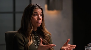 La Dra. Rhonda Patrick, una mujer de cabello castaño claro y largo, sentada con un suéter verde oscuro, gesticula con ambas manos abiertas en un fondo oscuro y desenfocado