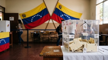 Urna transparente con sobres sobre una mesa de madera y dos banderas de Venezuela al fondo.