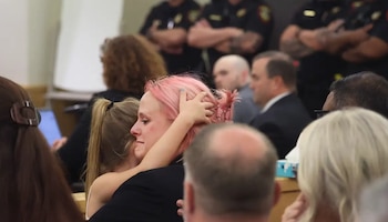 Una mujer con cabello rosa, con lágrimas en los ojos, es abrazada por una persona con cabello castaño, mientras una pequeña mano descansa sobre su cabeza en un evento concurrido