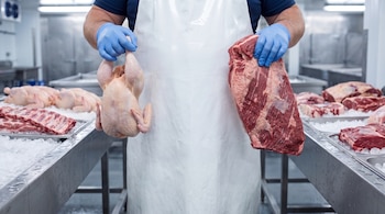 Hombre con delantal blanco y guantes azules, sujeta un pollo entero en mano izquierda y un corte de res en mano derecha; fondo de bandejas con carne y hielo.