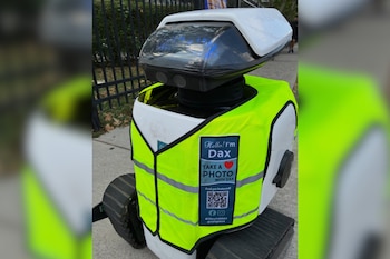 Primer plano de un robot blanco y negro con orugas, que lleva un chaleco amarillo reflectante con texto 'Hello! I'm Dax' y un código QR, en una acera