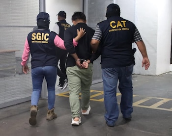 Dos agentes de investigación criminal con chalecos oscuros y pasamontañas escoltan por la espalda a un hombre esposado