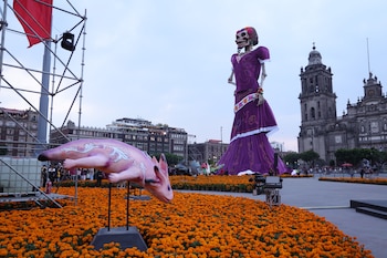 Una figura gigante de una calavera con vestido morado, una escultura de ajolote y flores de cempasúchil en una plaza frente a una catedral