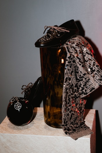 Un zapato negro con tachuelas descansa sobre un jarrón oscuro junto a una prenda de lentejuelas. Un objeto esférico negro con un charm plateado está en primer plano