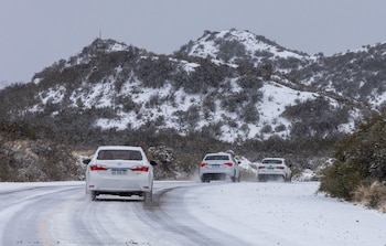 Tres automóviles blancos circulan por una carretera cubierta de nieve. Montañas con nieve y vegetación escasa se observan bajo un cielo gris