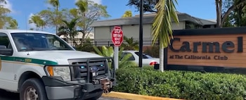 Camioneta Ford pickup blanca. Letrero de alto 'EXIT ONLY DO NOT ENTER'. Cartel 'Carmel at The California Club'. Palmeras, arbustos, edificio y coche blanco