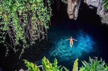 Vista aérea de una persona flotando en agua azul dentro de una cueva, rocas oscuras y vegetación verde colgando de la parte superior