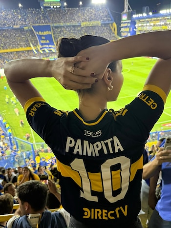 Vista trasera de Pampita en la Bombonera, con el pelo recogido y las manos en la cabeza, usando una camiseta de Boca Juniors negra y amarilla con "PAMPITA" y el número 10