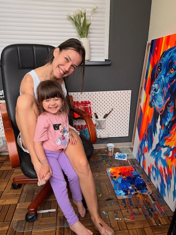 Una mujer y una niña en una silla negra junto a una paleta con pintura de colores y un cuadro de un perro azul en el suelo