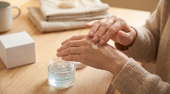 Primer plano de una persona aplicando crema transparente en el dorso de la mano izquierda con la derecha, junto a un tarro de hidrogel y una caja blanca sobre mesa de madera.