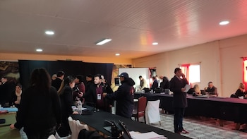 Interior de una sala con varias personas de pie y sentadas en mesas largas. Se ven computadoras, papeles, micrófonos y una pantalla de proyección