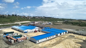 Vista aérea de campamentos mineros modulares blancos con techos azules, contenedores de carga, terreno en construcción y colinas verdes al fondo