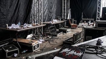 Backstage caótico con mesas plegables, botellas, papeles arrugados, cables desenrollados, cajas de equipo y una credencial de Hell & Heaven en el suelo.