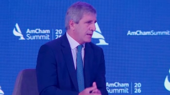 Luis Toto Caputo, un hombre de mediana edad con traje azul y corbata turquesa, habla en un evento con el logo de AmCham sobre un fondo azul