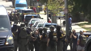 Calle con un grupo de agentes de seguridad uniformados, incluyendo algunos con "DEA" visible, junto a vehículos policiales y cinta de seguridad amarilla. Civiles y árboles
