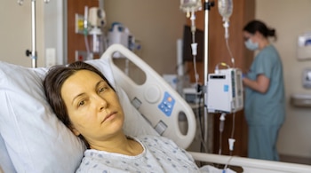 Mujer en cama de hospital con piel y ojos amarillentos. Cerca, un soporte para suero con bolsas y tubos. Al fondo, una enfermera con mascarilla.