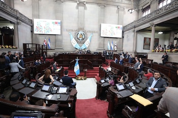 Interior del Palacio del Organismo Legislativo en Guatemala con diputados sentados y de pie, banderas de Guatemala y pantallas mostrando resultados de votación