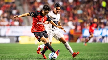 Dos jugadores disputan un balón en una cancha de césped; uno lleva uniforme rojo con negro y el otro viste completamente de blanco.