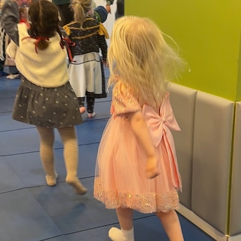 Niña rubia de espaldas en un vestido rosa brillante con un lazo grande, caminando en un espacio interior con suelo azul y pared verde, con otra niña cerca