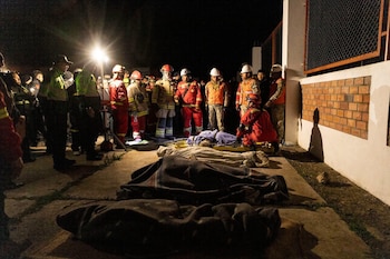 Grupo de rescatistas y personas con uniformes de trabajo rodean cuerpos cubiertos con mantas y una camilla en el suelo. La escena está iluminada artificialmente de noche