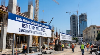 Vista de un sitio de construcción activo con edificios en varias etapas, grúas, trabajadores con chalecos y cascos de seguridad, y pancartas informativas.